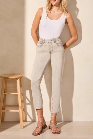 A woman standing next to a wooden stool, wearing a white sleeveless top and slim crop jeans in a shadowmist color.