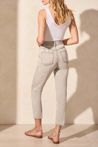 A woman standing next to a wooden stool, wearing a white sleeveless top and slim crop jeans in a shadowmist color.