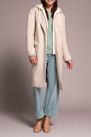 Person wearing a long beige coat with a hood, light blue jeans, and brown shoes on a plain background.