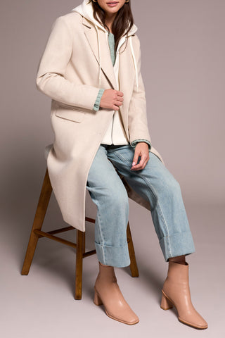 Woman wearing a long cream coat, blue jeans, and beige boots sitting on a wooden stool.