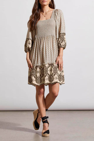 A woman wearing in a Tribal Wear two-ways embroidered dress in French Oak with square neckline and tie-closure back, paired with tan ankle strap sandals.