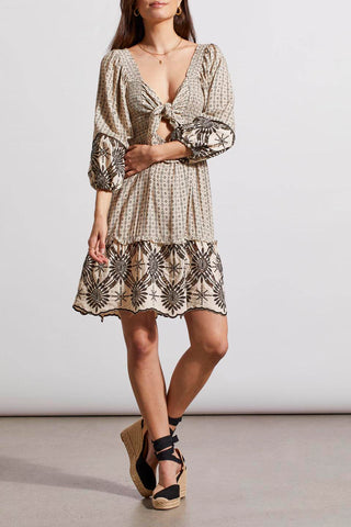 A woman wearing in a Tribal Wear two-ways embroidered dress in French Oak with square neckline and tie-closure back, paired with tan ankle strap sandals.