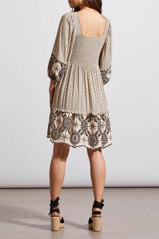 A woman wearing in a Tribal Wear two-ways embroidered dress in French Oak with square neckline and tie-closure back, paired with tan ankle strap sandals.