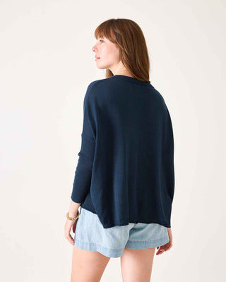 Woman wearing a navy sweater over a light blue shirt with denim shorts on a white background. Mersea Catalina Cotton Crewneck Sweater in Navy