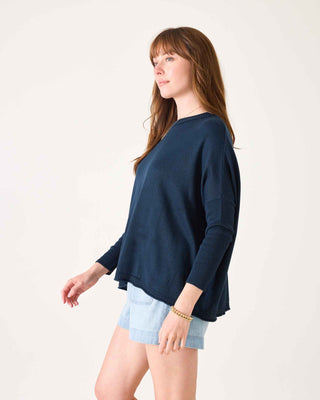 Woman wearing a navy blue sweater and light blue shorts on a white background. Mersea Catalina Cotton Crewneck Sweater in Navy