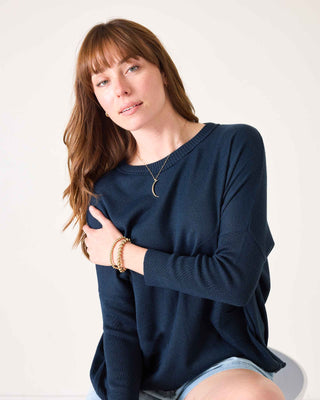 Woman wearing a navy blue sweater and denim shorts on a white background. Mersea Catalina Cotton Crewneck Sweater in Navy