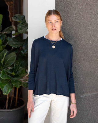 Woman wearing a navy blue sweater and white pants standing against a gray wall with a plant to the left. Mersea Catalina Cotton Crewneck Sweater in Navy