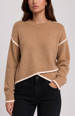 A woman wearing a cropped, long-sleeve knit sweater in camel beige with contrasting cream trim along the shoulders, hem, and cuffs, paired with black jeans against a light gray studio background. Liverpool Long Sleeve Camel Sweater with Cream Tulip Hem