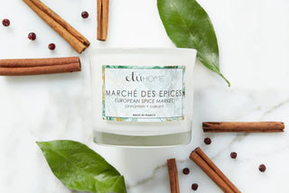 A clear glass candle jar with white soy wax, labeled 'Marché des Épices', surrounded by cinnamon sticks and green leaves on a white marble surface. ETU