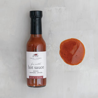 A bottle of Fire Roasted hot Sauce with a small amount of sauce visible in front of it on a white surface, finch and fennel